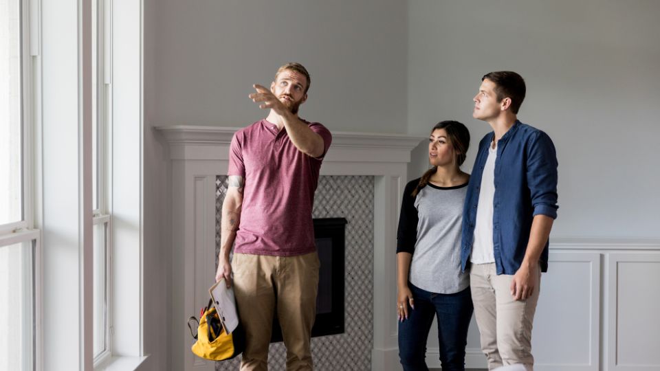 Two renters stand in front of a fireplace while a repairman, holding a toolbox and clipboard, points to the window and speaks to them.